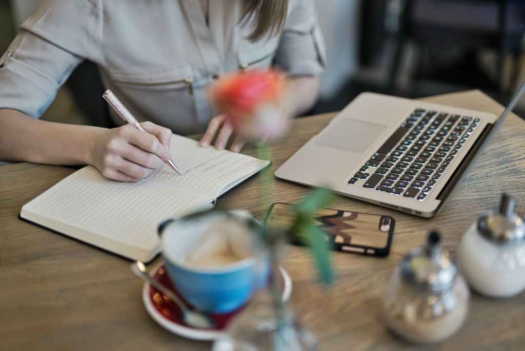 Woman writing in notebook with coffee and computer and phone